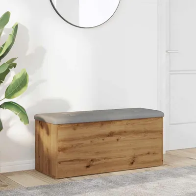 A wooden storage bench with a gray upholstered top is placed in a hallway next to a large potted plant and a mirror on the wall.