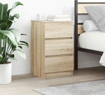 A bedside table with three drawers is placed next to a bed, with a large potted plant on the left.