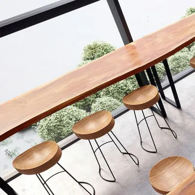 A long, live-edge wooden bar table with several wooden stools in front of a large window with greenery outside.