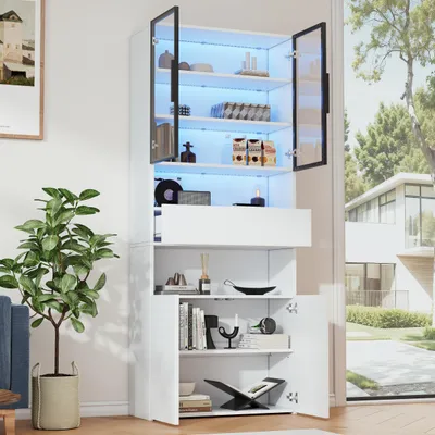 A white display cabinet with glass doors and LED lighting, filled with various decorative items and books. The cabinet is placed in a room with a plant and a window showing a modern house and garden outside.