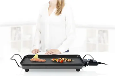 A person is standing behind a table with an electric grill on it. The grill has food on it.