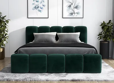 A green upholstered bed with two pillows and a dark gray blanket is shown in a bedroom setting. Two framed pictures hang on the wall behind the bed, and two potted plants are on either side.
