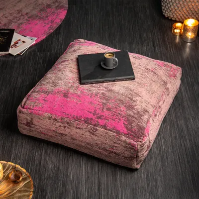 A square floor cushion with a pink and brown abstract pattern sits on a dark wooden floor. A small book and a cup of coffee are placed on top of the cushion. In the background, there are books, candles, and a woven basket.