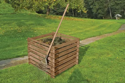 A wooden compost bin with a rake in a grassy yard with trees in the background.