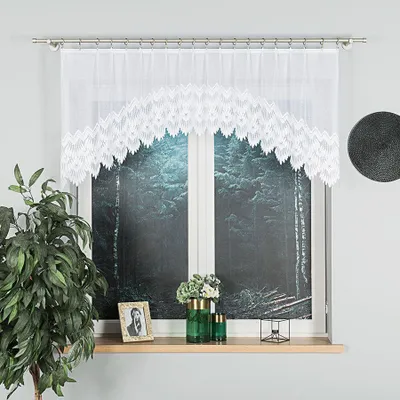 A white lace curtain with a geometric pattern is shown hanging over a window with a forest scene printed on it. A plant and some decorative items are on the windowsill.