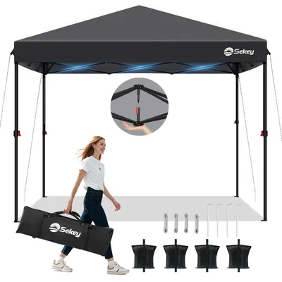 A woman is walking with a carrying bag for a pop-up canopy tent. The tent is set up in the background.