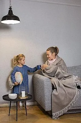 A woman and a child are in a living room setting. The woman is sitting on a couch wrapped in a blanket, and the child is standing next to her, holding a pillow and reaching out to the woman.