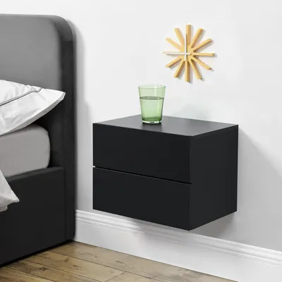 A black floating nightstand with two drawers is mounted on a white wall next to a bed. A glass of water and a wooden clock are on top of the nightstand.