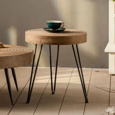 A close-up shot of a round wooden side table with a dark green teacup and saucer on top, set against a neutral gray wall and a wooden floor.