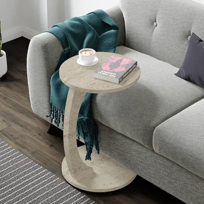 A C-shaped side table with a book and coffee cup on it, placed next to a sofa with a teal blanket draped over it.