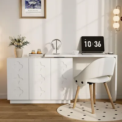 A white desk with a laptop, headphones, and a vase of flowers on top, next to a white cabinet with decorative doors. A white chair is placed in front of the desk.