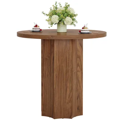 A round wooden dining table with a vase of flowers and two small cakes on top, set against a white background.