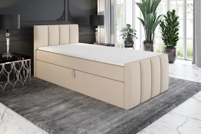 A beige boxspring bed with a mattress and topper is shown in a bedroom setting with plants and lamps.