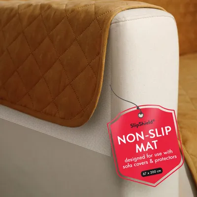 A close-up of a tan quilted sofa protector on a light-colored sofa armrest. A red tag with product information hangs from the protector.