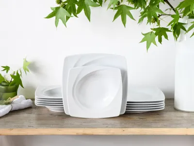 A stack of white square plates and bowls are arranged on a wooden shelf, with green leaves and a white vase in the background.