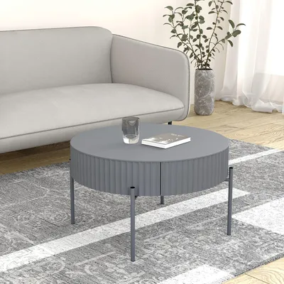A round, gray coffee table with a fluted design and metal legs sits on a patterned rug in a living room setting. A sofa is visible in the background, along with a vase of plants and sheer curtains.