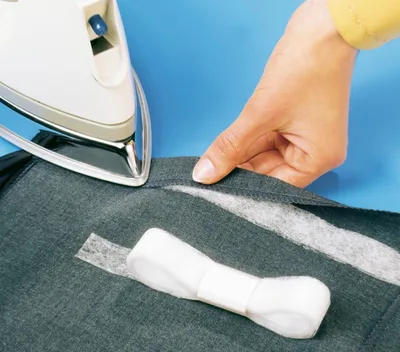 A person is using an iron to attach a strip of fusible interfacing to a piece of gray fabric.