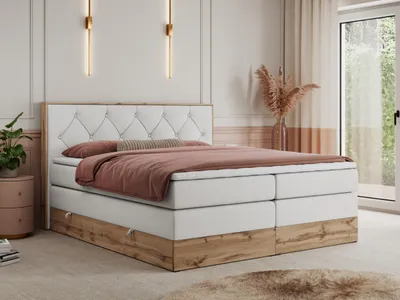 A bedroom scene featuring a king-size boxspring bed with a tufted headboard and a wooden base. The bed is dressed with pink bedding and a bolster pillow. A bedside table with a plant is visible to the left, and a vase with dried flowers is on the right.