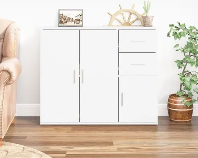A white sideboard with two doors and two drawers, placed against a white wall. A sofa is visible on the left, and a potted plant is on the right. Decorative items are on top of the sideboard.