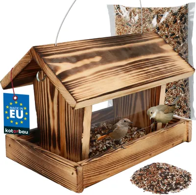 A wooden bird feeder filled with birdseed, with two small birds perched on it. A bag of birdseed and a pile of birdseed are also visible.