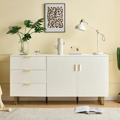 A white sideboard with gold accents, a plant, a lamp, and artwork on a light yellow wall.