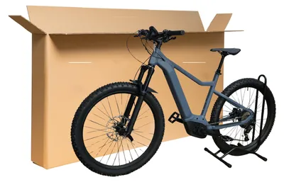 An electric bicycle is shown in front of a large cardboard box, with a bike stand to the right.
