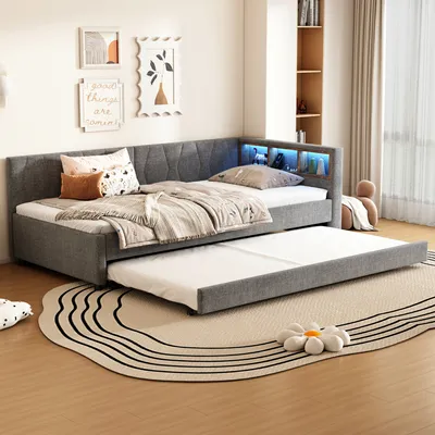 A modern bedroom with a gray upholstered daybed with a pull-out trundle bed. The daybed has built-in LED lighting in the headboard and is adorned with decorative pillows and blankets. A round rug with a wavy pattern is placed in front of the bed.