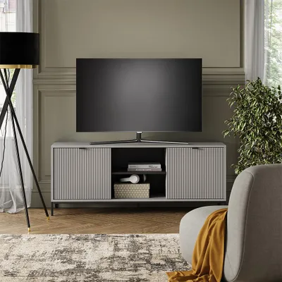 A living room scene featuring a TV stand with a television on top, a floor lamp, a plant, and a chair with a throw blanket.