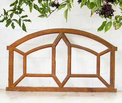 A rusty cast iron window frame is displayed against a white brick wall with greenery hanging above.