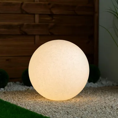 A white spherical garden light sits on a bed of white gravel, with a wooden fence and greenery in the background.
