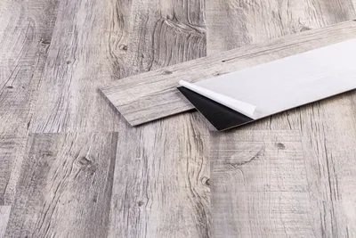 Close-up of self-adhesive vinyl planks with a wood grain texture, showing the peel-back adhesive backing of one plank.
