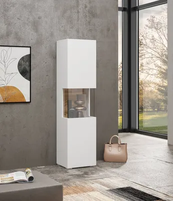A modern white vitrine cabinet with glass accents is placed against a textured gray wall, next to a large window with a view of trees. A beige handbag sits on the floor nearby.