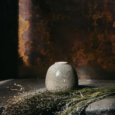 A speckled vase sits on a dark surface with dried grass in the foreground, against a textured, dark brown background.