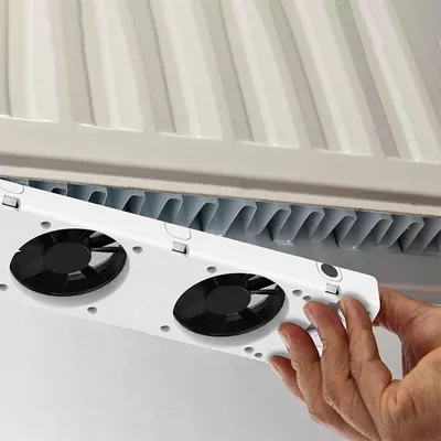 A hand is holding a white device with two black fans, which is being attached to a radiator.