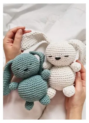 Two crocheted bunnies, one eucalyptus green and one white, are held by a person's hands on a white background.
