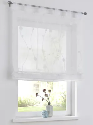 A white sheer curtain with a subtle embroidered pattern hangs over a window, with a vase of dried flowers and a small decorative object on the windowsill below.