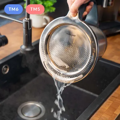 A person is holding a stainless steel sieve over a black sink, with water draining through it. Steam is rising from the sink.