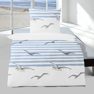 A duvet cover and pillowcase set with a blue and white striped pattern and seagull illustrations, displayed on a bed in a room with a window overlooking a beach.