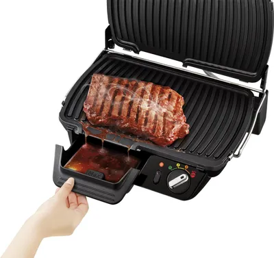 A hand is shown pulling out the drip tray of a black electric grill with a steak cooking on it.