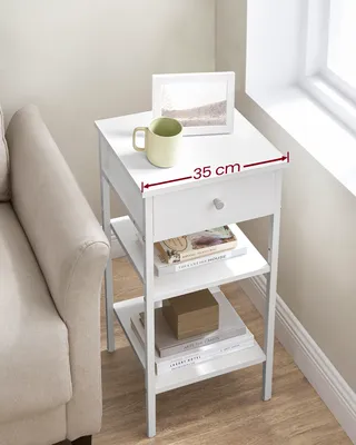 A white bedside table with a drawer and two shelves is shown next to a couch, near a window. The table has a mug, a framed picture, and books on it.