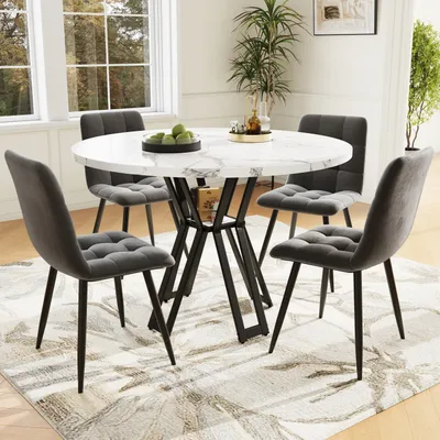 A dining set with a round marble-top table and four gray upholstered chairs, placed on a patterned rug in a well-lit room with windows.