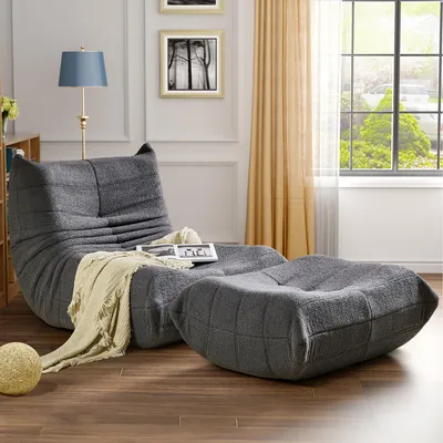 A gray beanbag chair and ottoman are set up in a living room with a lamp and artwork on the wall, next to a window with curtains.