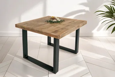 A square wooden table with black metal legs is shown in a well-lit room with a tiled floor and a plant in the background.