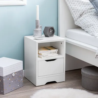 A white nightstand with a drawer and shelf, decorated with a candle, a small clock, and books, placed next to a bed in a bedroom.