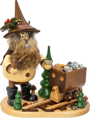 Ein Erzwichtel mit spitzem Hut und buschigem Bart steht neben einem Holzwagen voller bunter Steine auf einer Holzplatte dekoriert mit kleinen Tannenbäumen und hält einen Hammer in der Hand während er freundlich lächelt