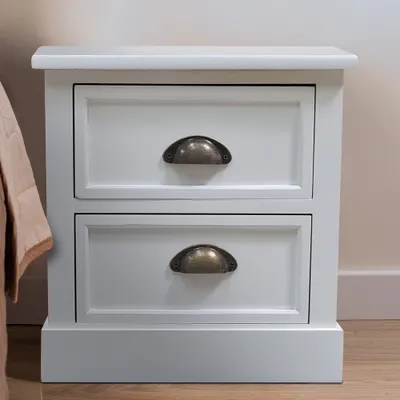 A white wooden nightstand with two drawers and bronze handles sits next to a bed with a beige quilted throw.
