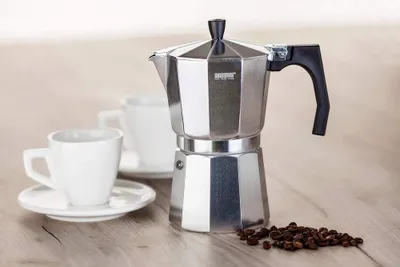 A coffee maker with two cups and coffee beans on a wooden surface.