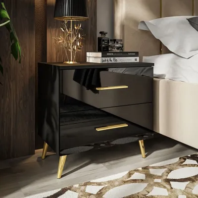 A modern black high-gloss nightstand with gold legs and gold drawer pulls, placed next to a bed. A lamp and books are on top of the nightstand.