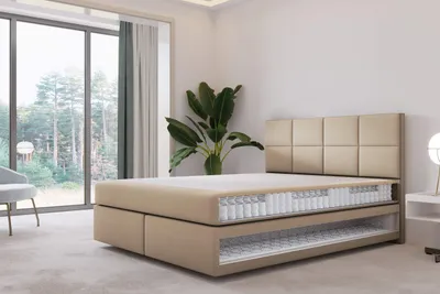 A cutaway view of a beige box spring bed with a headboard in a well-lit room with large windows.