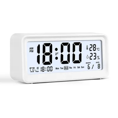 A white digital alarm clock is shown against a white background. The clock displays the time, date, temperature, and humidity.